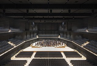 Interimsspielstätte Gasteig München Foto der Interimsspielstätte Gasteig in München. Zu sehen ist der bestuhlte Bühnenraum mit Orchester.
