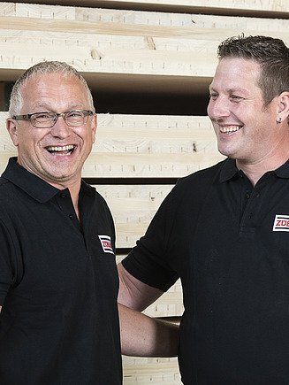 Teamgeist bei ZÜBLIN Timber Foto von zwei Männern die lachen. Im Hintergrund ist ein LENO-Brettsperrholzstapel zu sehn.